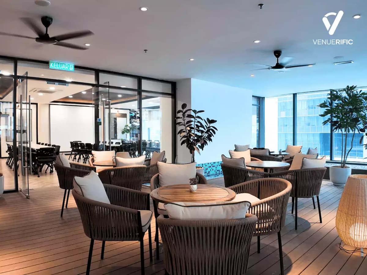 Modern lounge area with woven chairs, round wooden tables, ceiling fans, and glass doors leading to a meeting room.