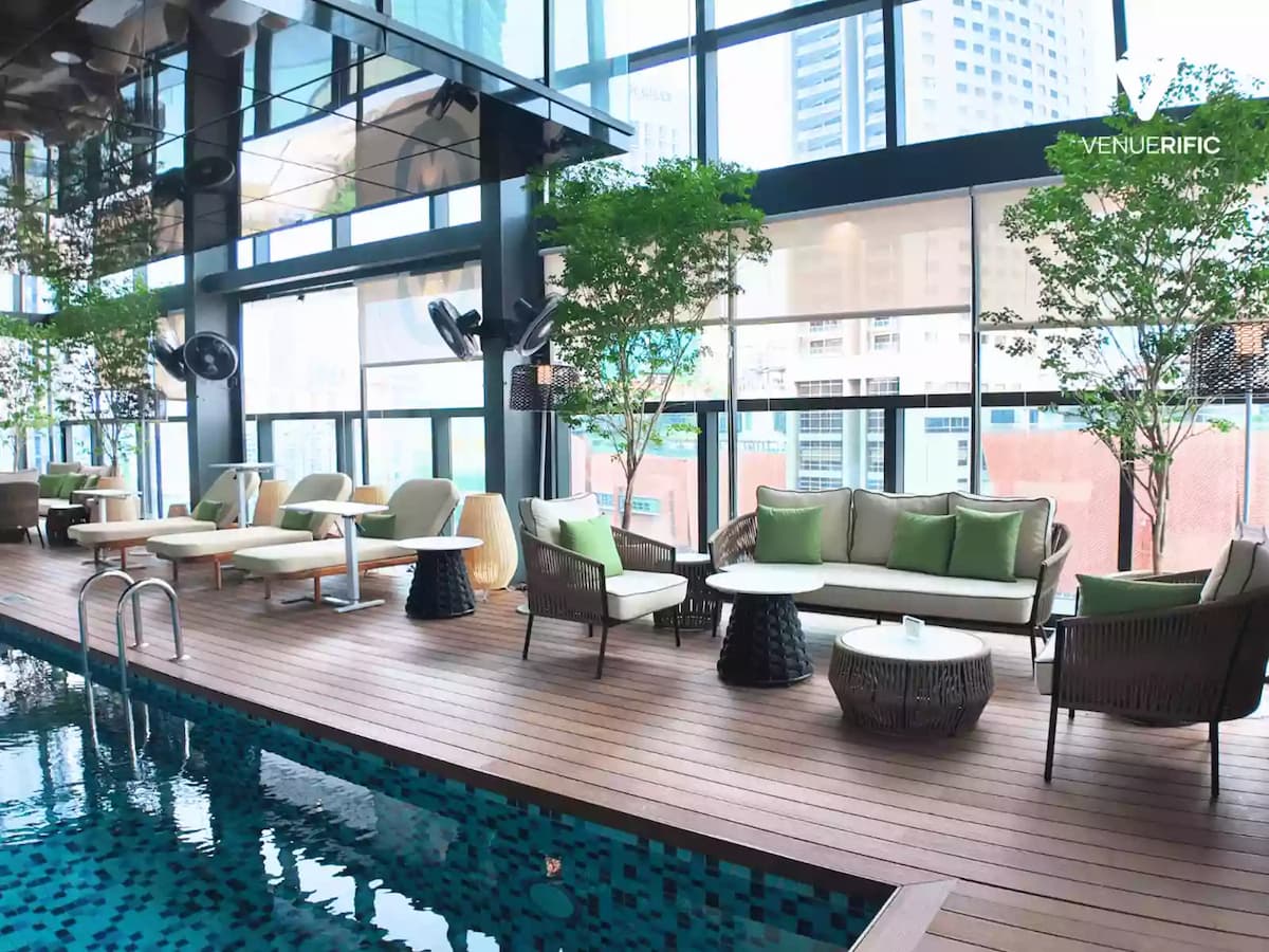 Indoor poolside lounge with cushioned seating, wooden deck flooring, and large windows overlooking the city.
