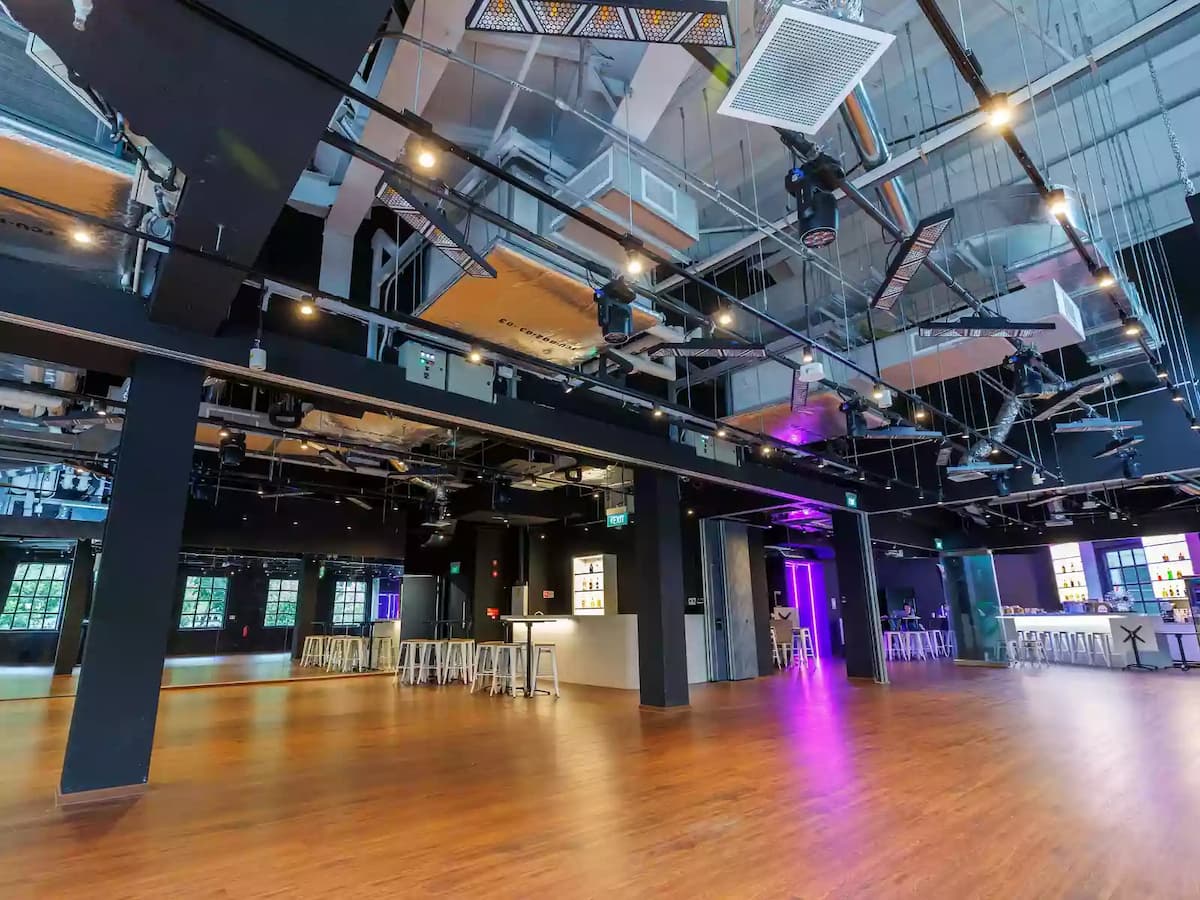Industrial-style event hall with wooden flooring, exposed ceiling rigging, bar counter, and open layout.