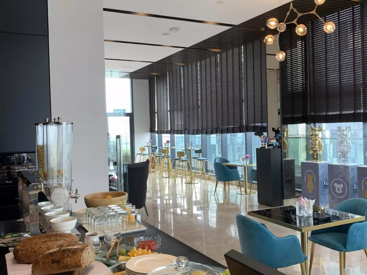 Modern lounge dining area with buffet counter, teal chairs, pendant lights, and floor-to-ceiling windows with city views.