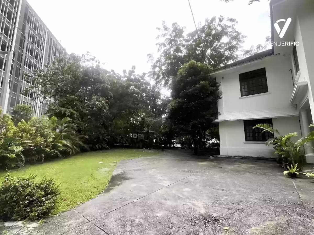 Outdoor courtyard with lawn and paved area beside a white heritage-style building surrounded by trees and greenery.