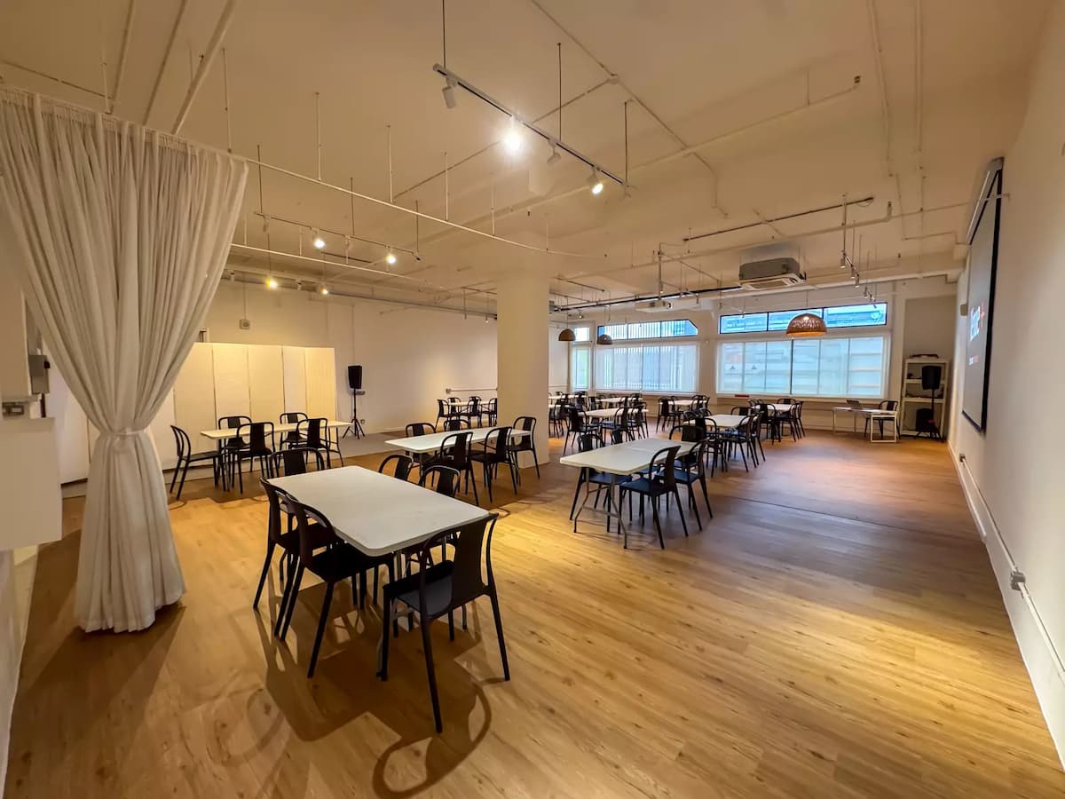 Bright event space with white walls, wooden floors, communal tables, and rattan pendant lighting for workshops or gatherings.