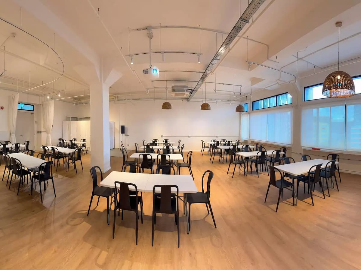 Spacious event hall with communal tables, wooden floors, natural light, and rattan pendant lighting for gatherings or workshops.