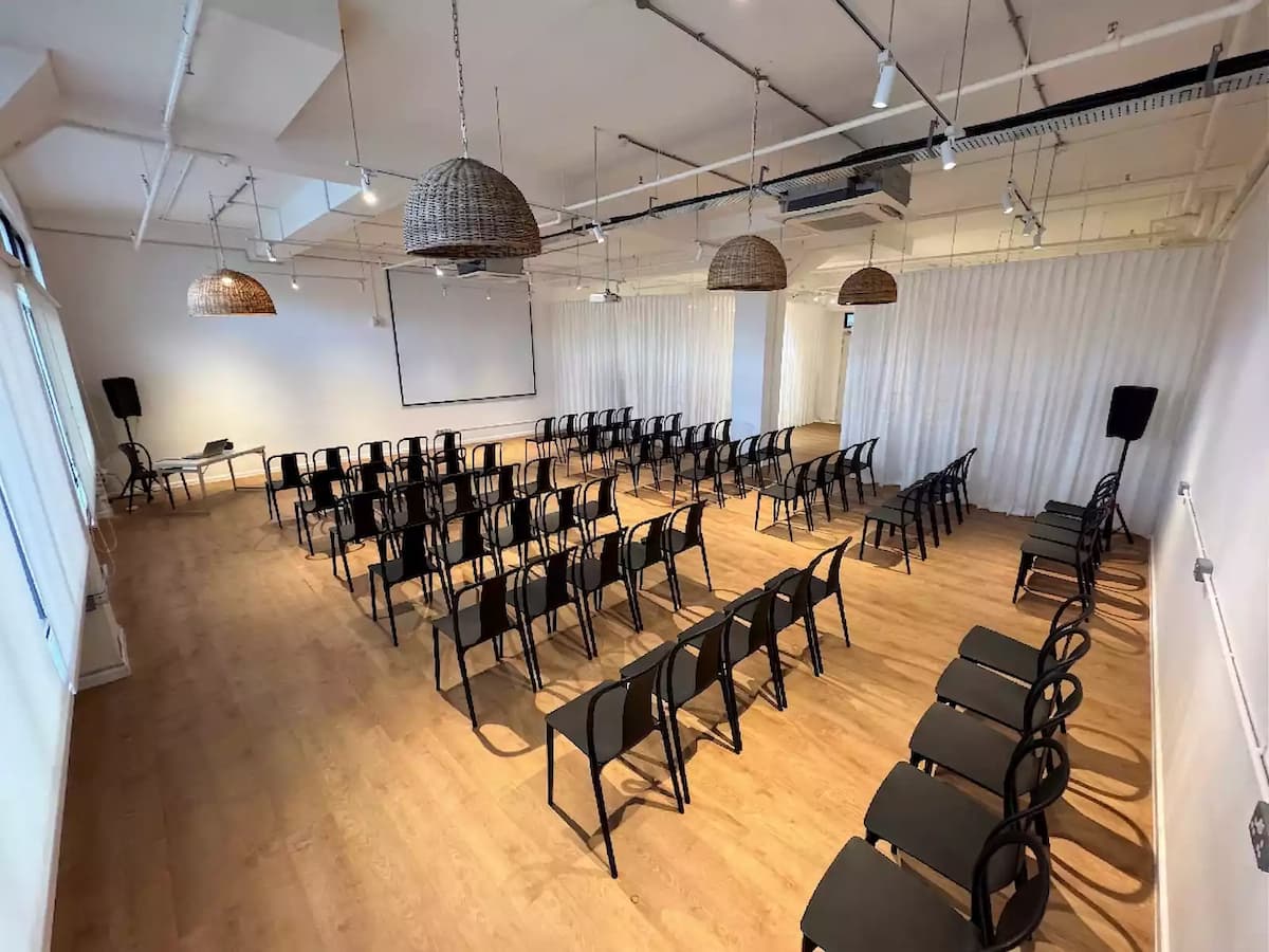 Bright event space with rows of chairs, projector screen, wooden floors, and rattan pendant lighting for presentations or workshops.