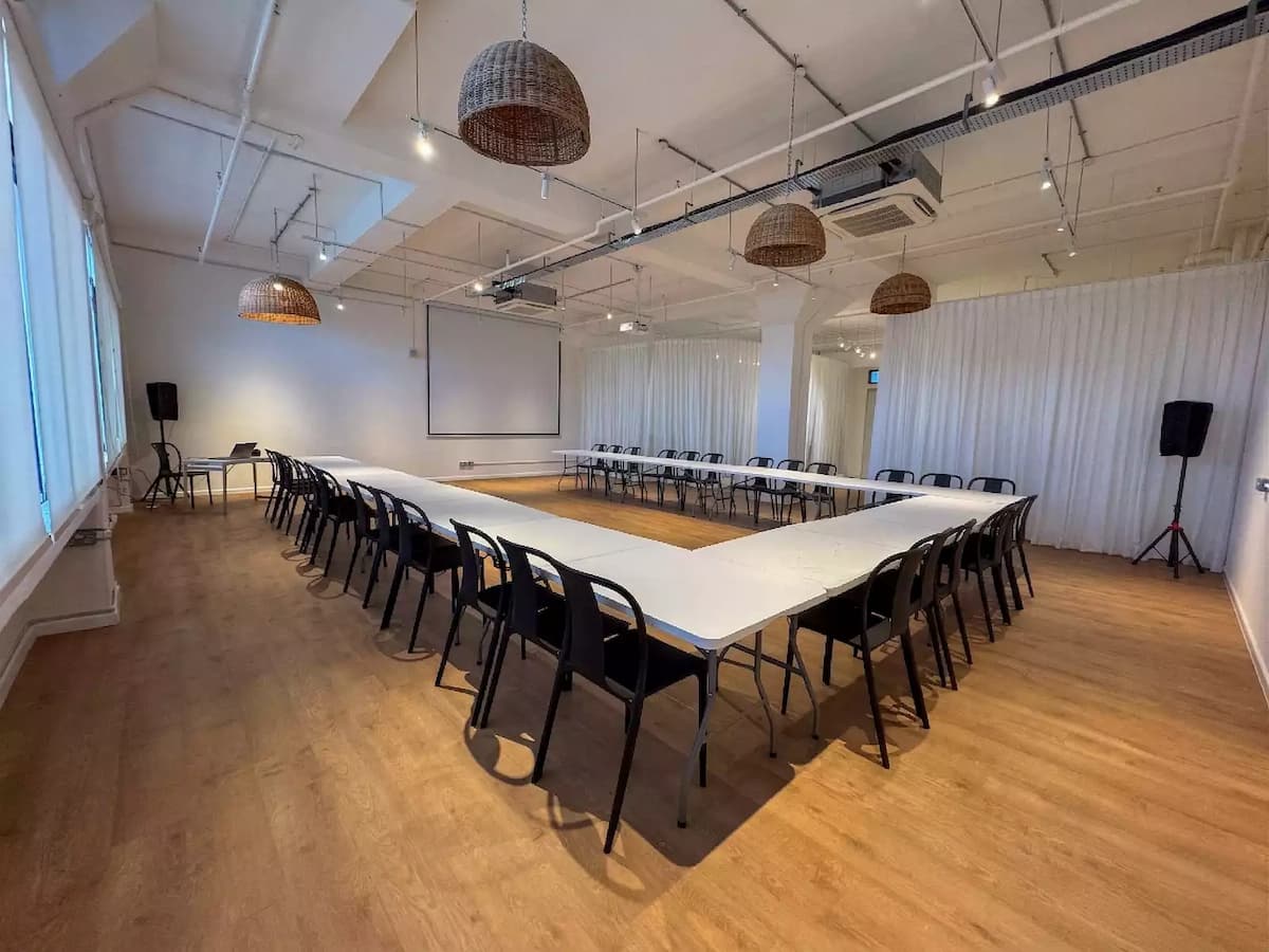 Bright event space with U-shaped table setup, white walls, wooden floors, and rattan pendant lighting for meetings or workshops.