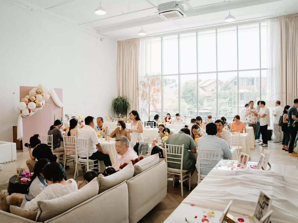 birthday celebration in a bright minimalist studio with guests seated at white chiavari chair tables and a pastel balloon backdrop