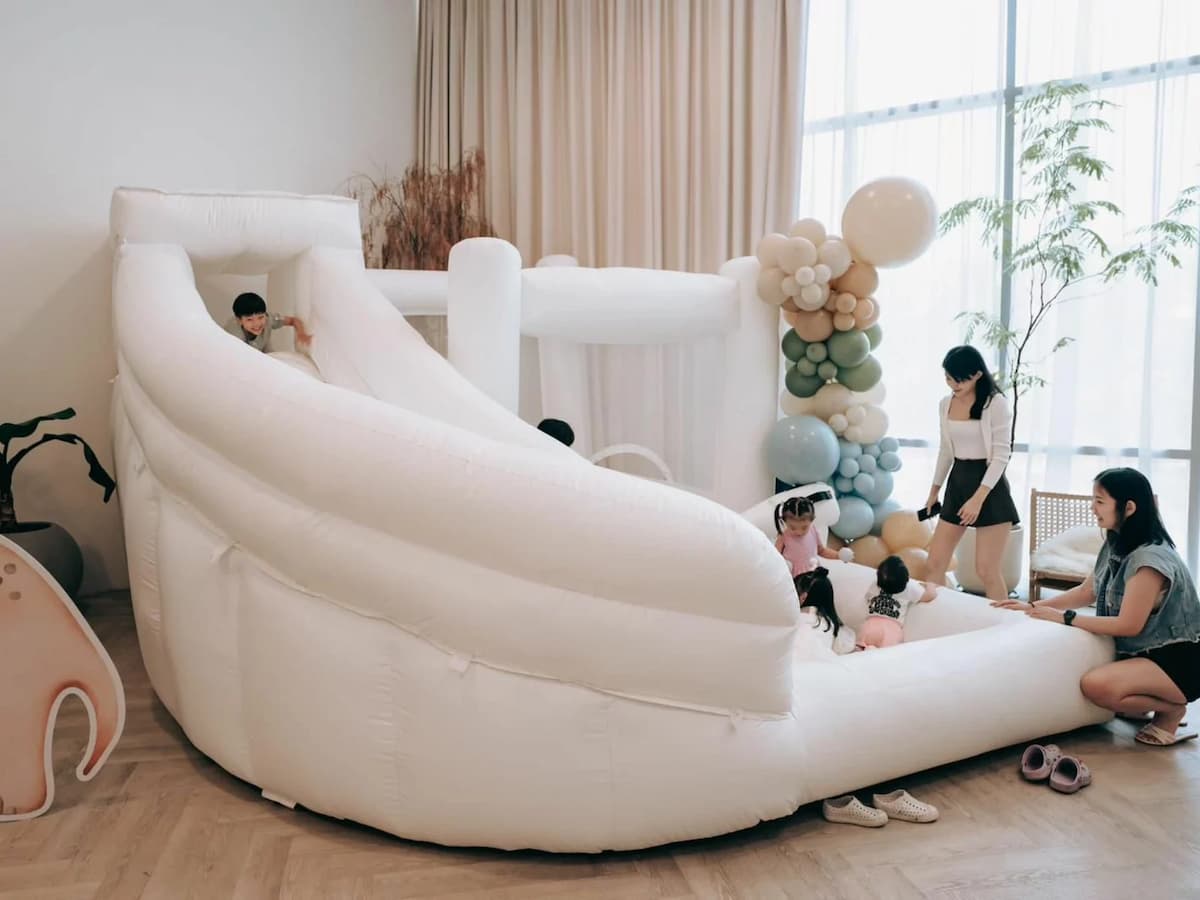 Children playing on a white inflatable bounce castle with slide in a bright studio, decorated with sage and cream balloon garland — ideal for kids' birthday parties