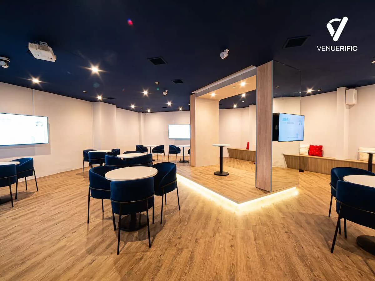 Modern event space with blue ceiling, round tables and chairs, presentation screens, and a central wooden structure with ambient lighting.