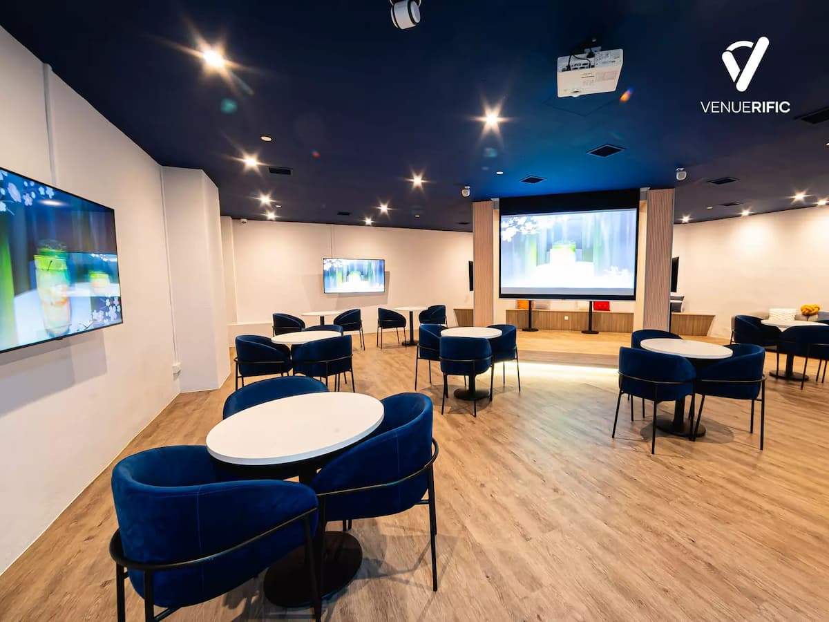 Modern event space with round tables, blue chairs, wall screens, and a large presentation screen at the front.