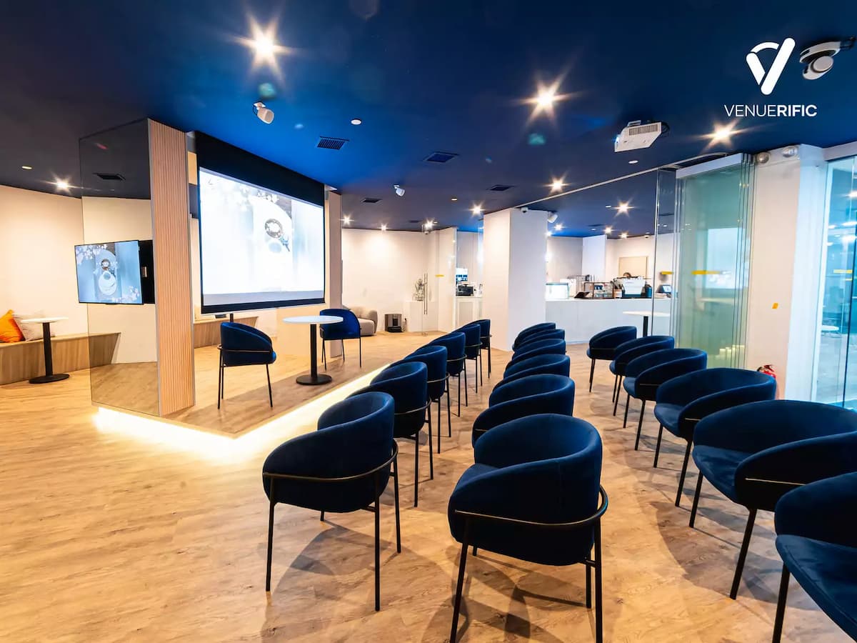 Modern presentation space with rows of blue chairs, large screen, and stage area with table seating.
