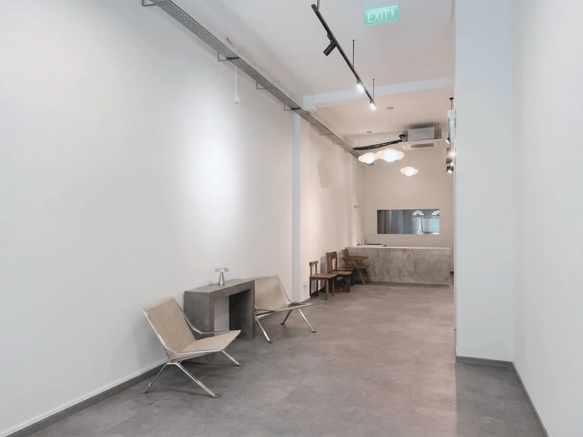 Minimalist event space with white walls, concrete finishes, and simple seating along a long, open layout.