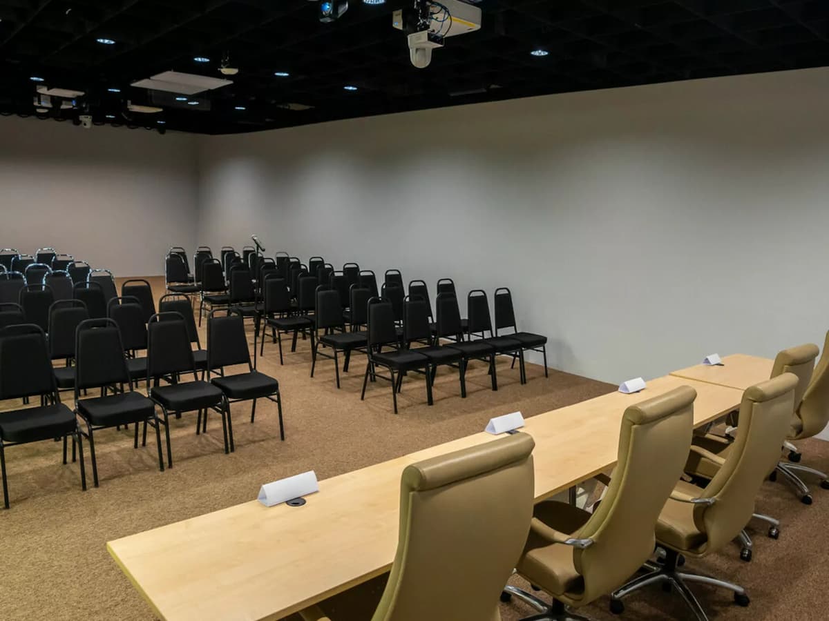 Seminar room with rows of black chairs facing a panel table and ceiling-mounted projector for presentations.