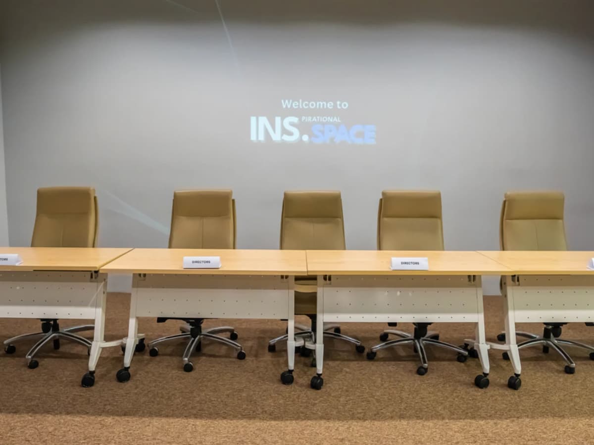 Conference panel setup with long meeting tables, office chairs, and a presentation screen on a minimalist wall.