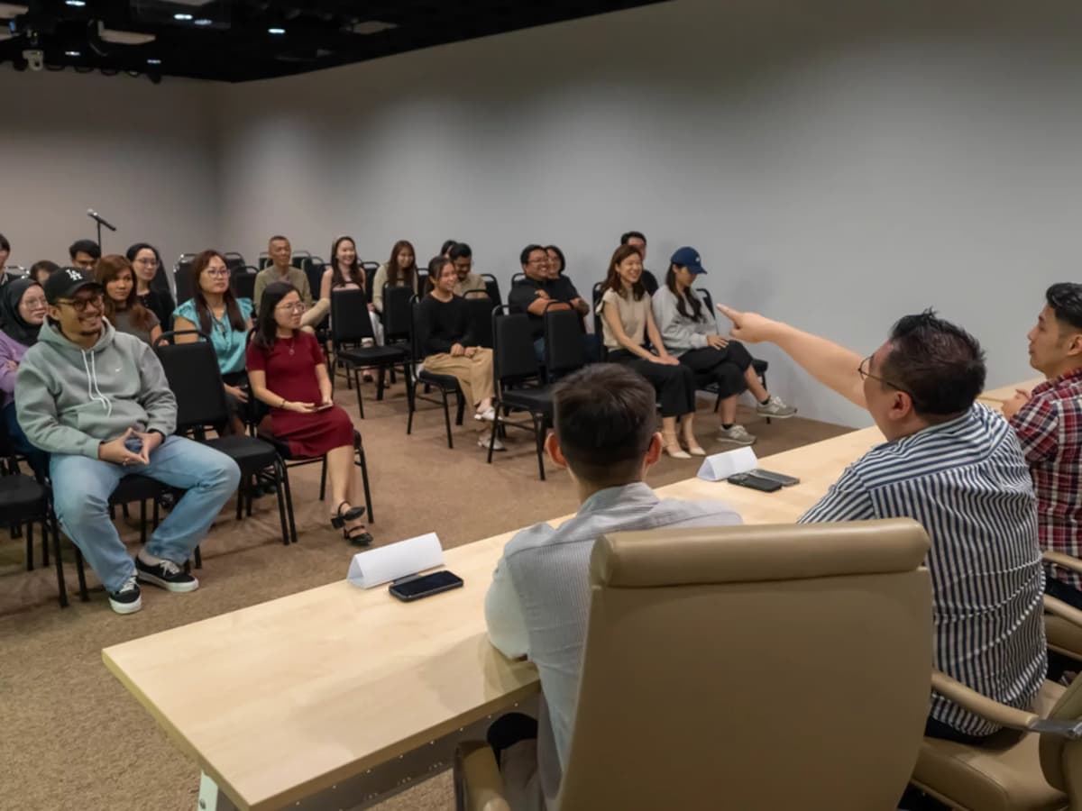 Speaker addressing an audience in a seminar-style room with seated attendees facing a panel table.