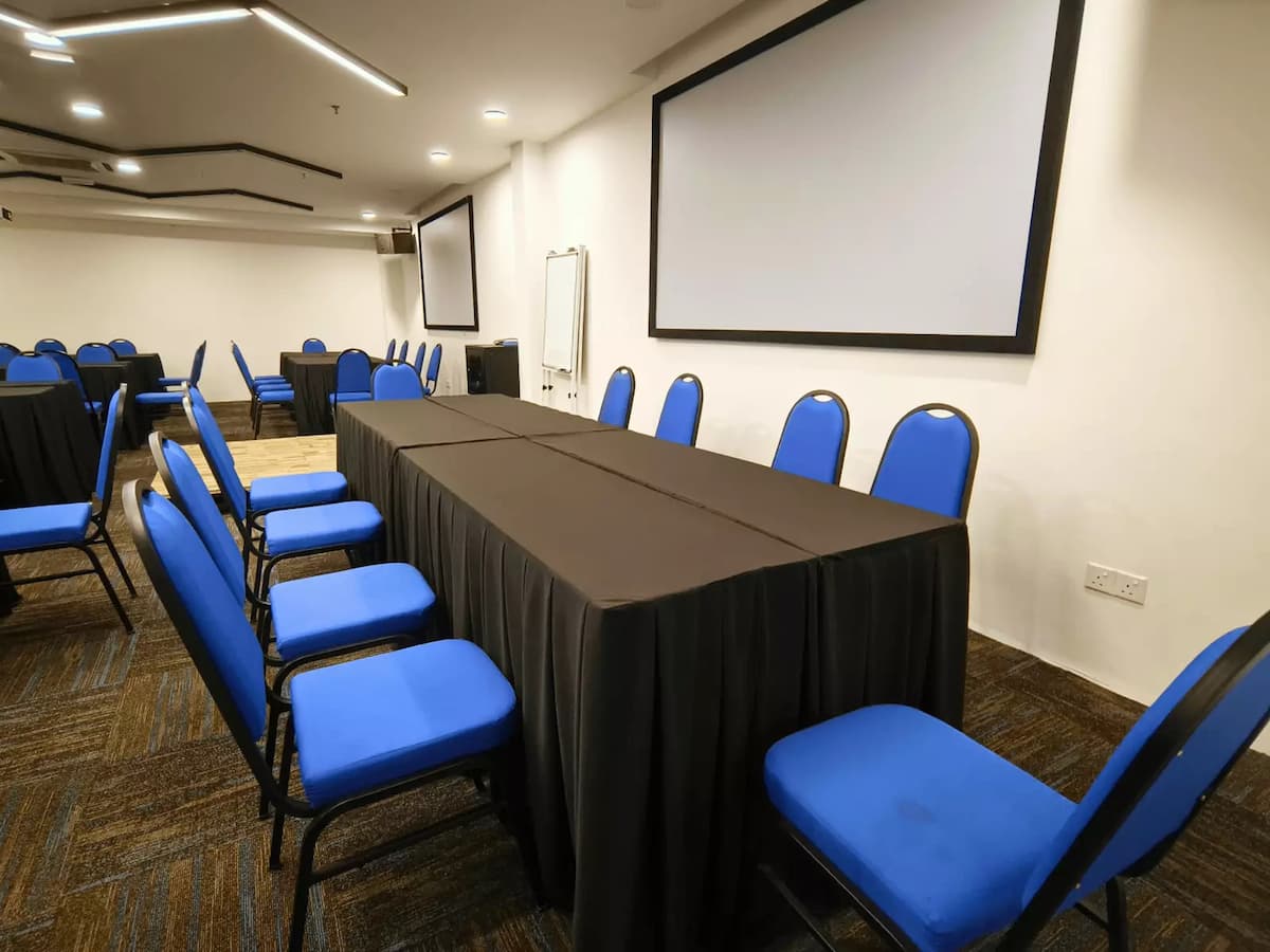 A professional room featuring blue cushioned chairs, long tables with black covers and a large projection screen