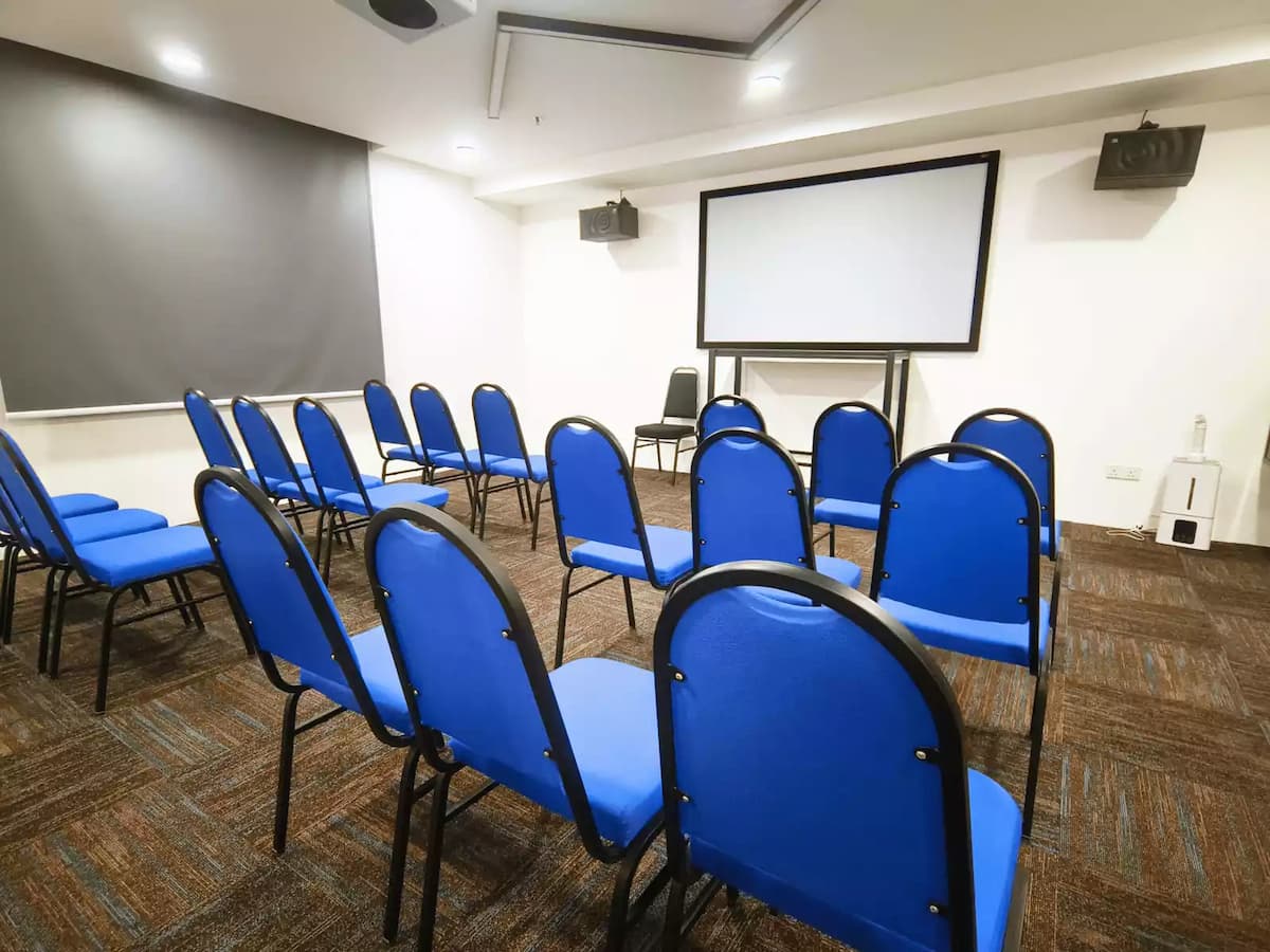 A well-lit seminar room featuring rows of blue cushioned chairs facing a projector screen