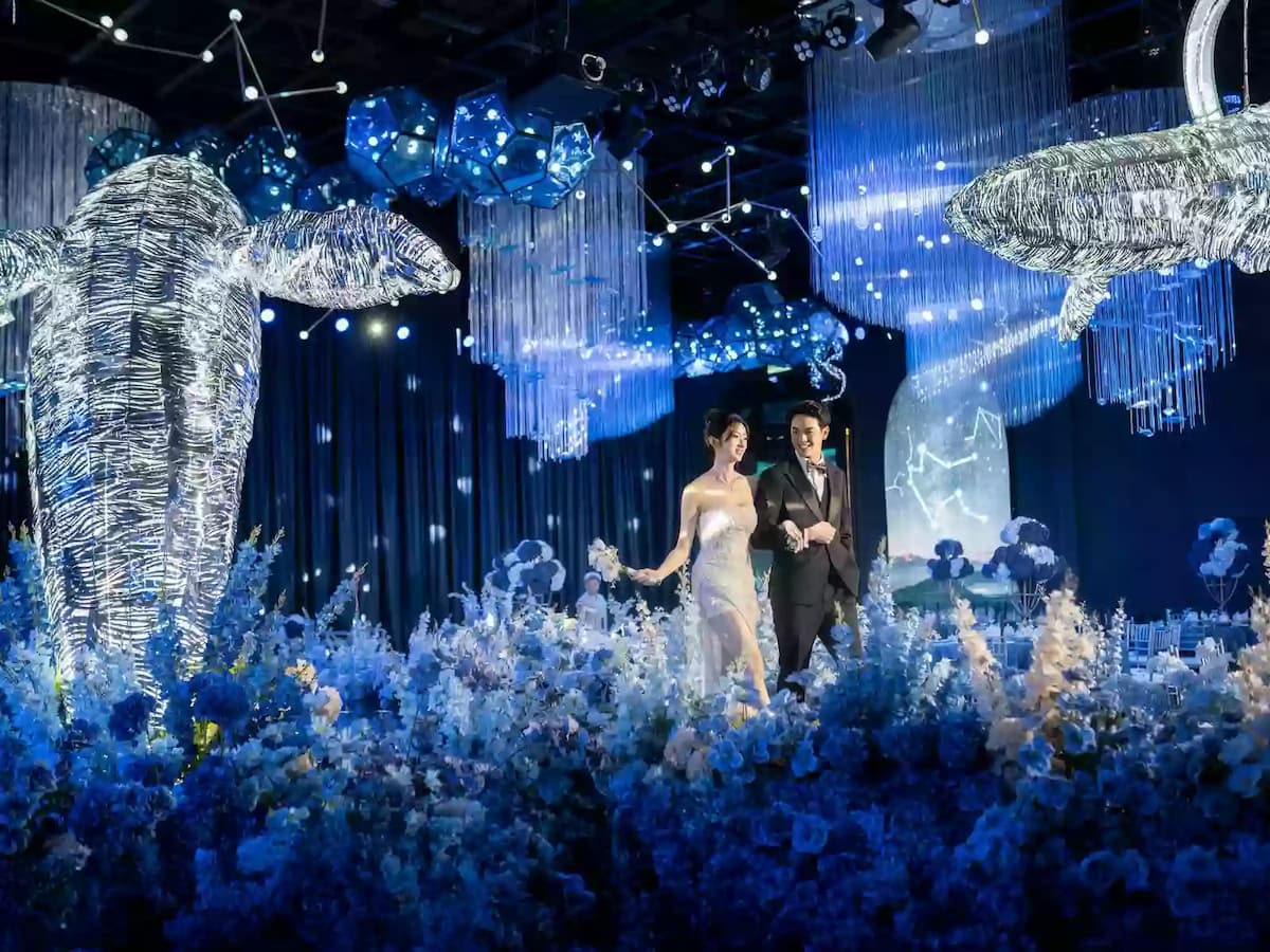 Elegant wedding stage with bride and groom walking through a floral aisle beneath illuminated whale sculptures and chandeliers.