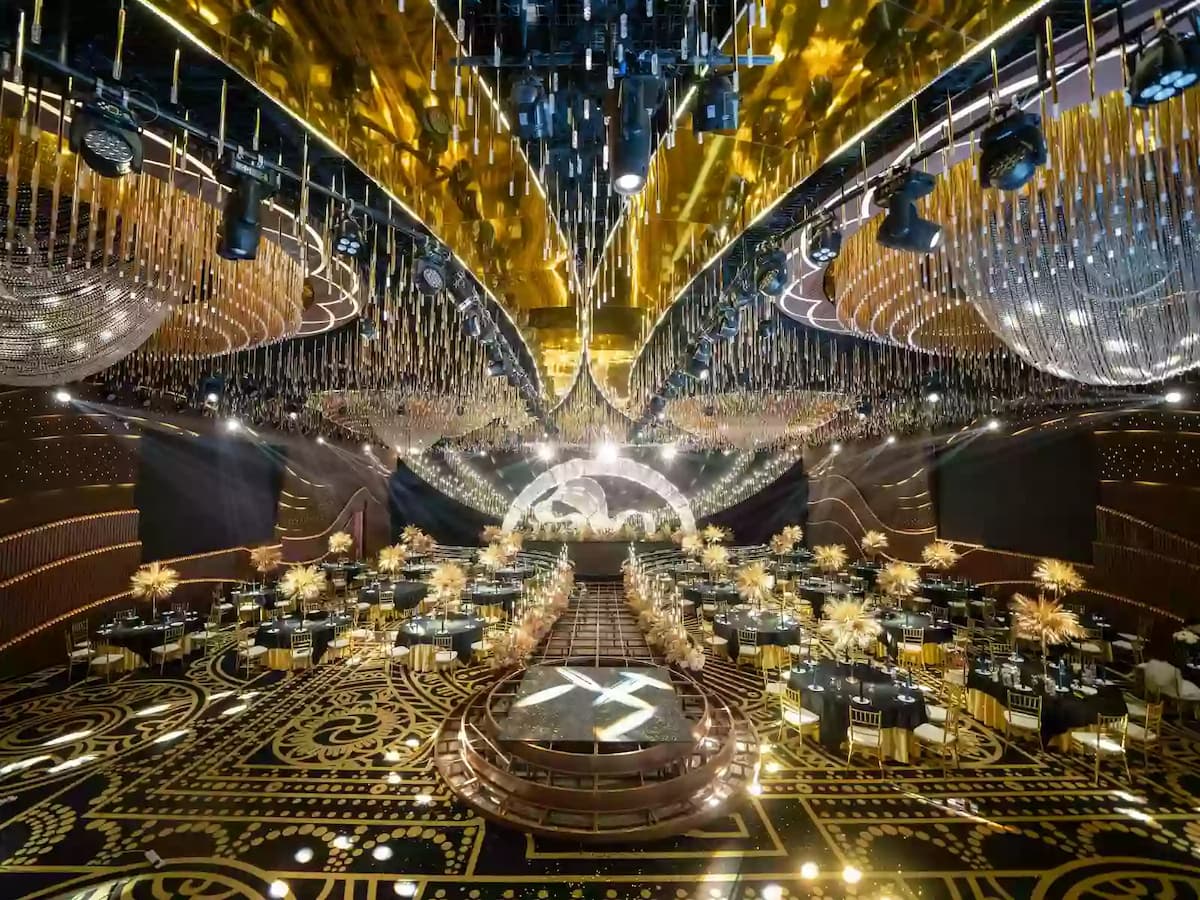 Lavish gold-themed ballroom with grand stage, cascading chandeliers, ornate patterned flooring, and banquet tables arranged around a central aisle.