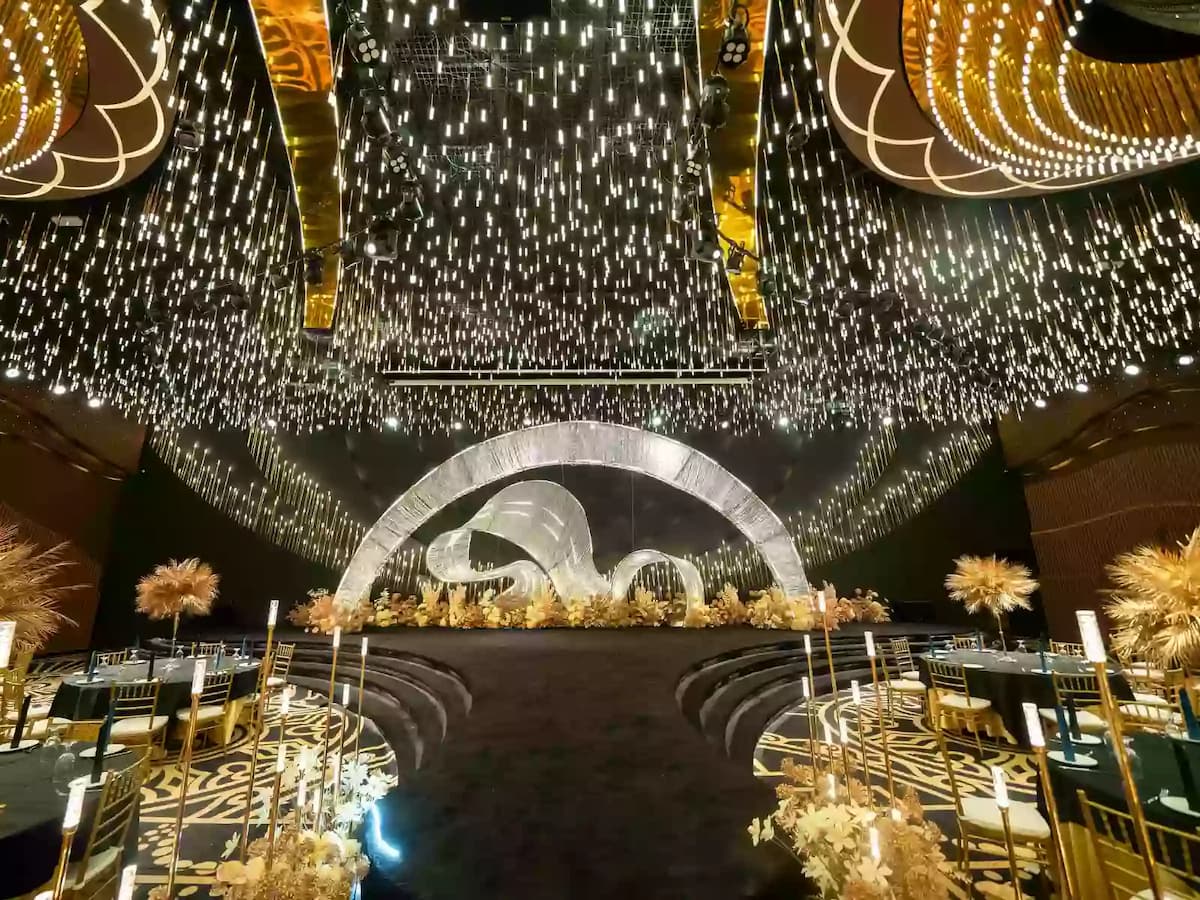 Grand wedding hall with sweeping stage, gold decor, cascading ceiling lights, and banquet tables arranged around a central aisle.