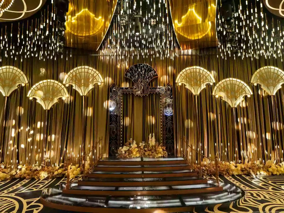 Luxurious gold-themed wedding stage with grand staircase, ornate arches, cascading lights, and floral arrangements.