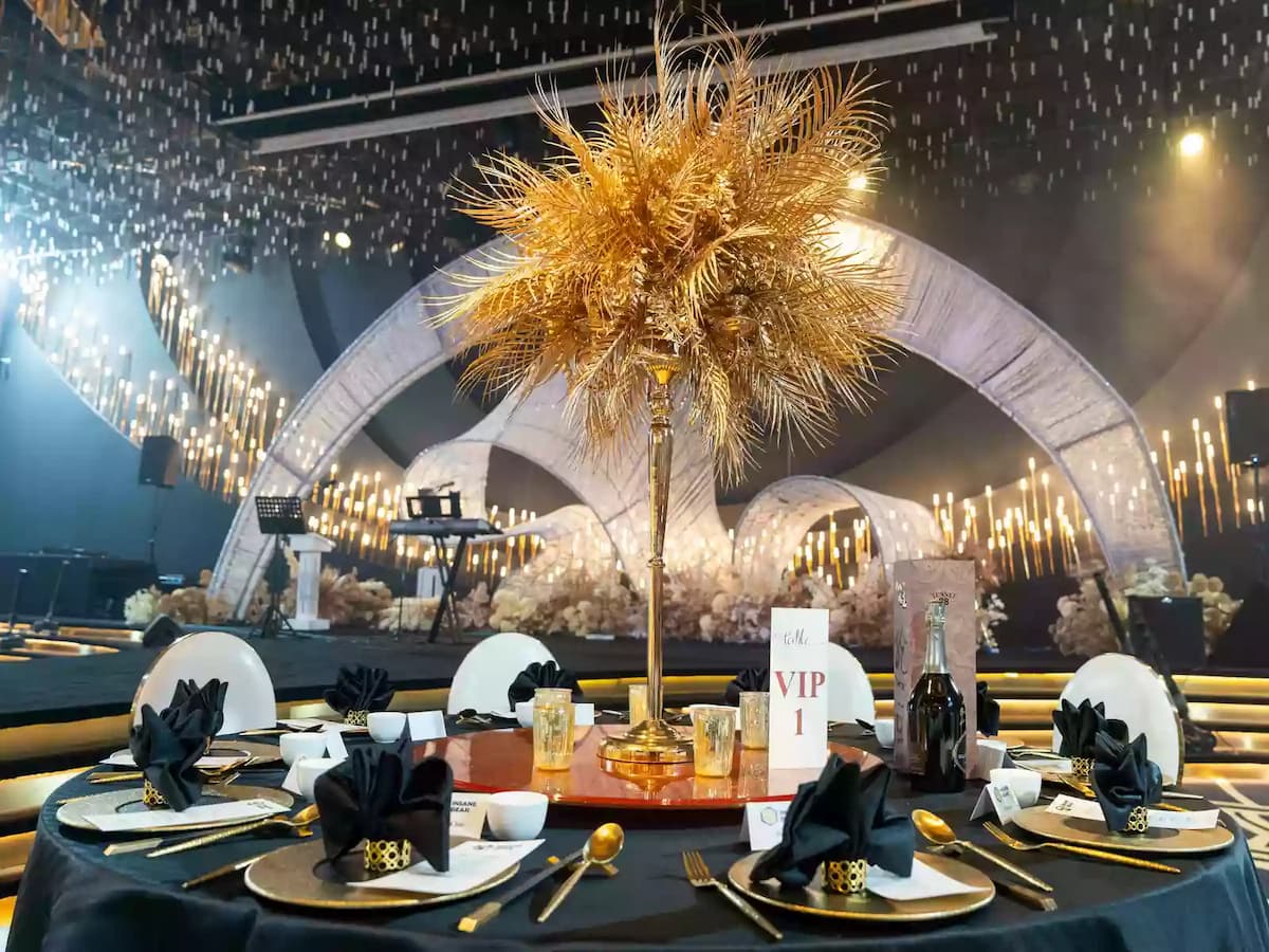 Elegant banquet hall with gold chairs, black table settings, and tall golden feather centerpieces under warm lighting.