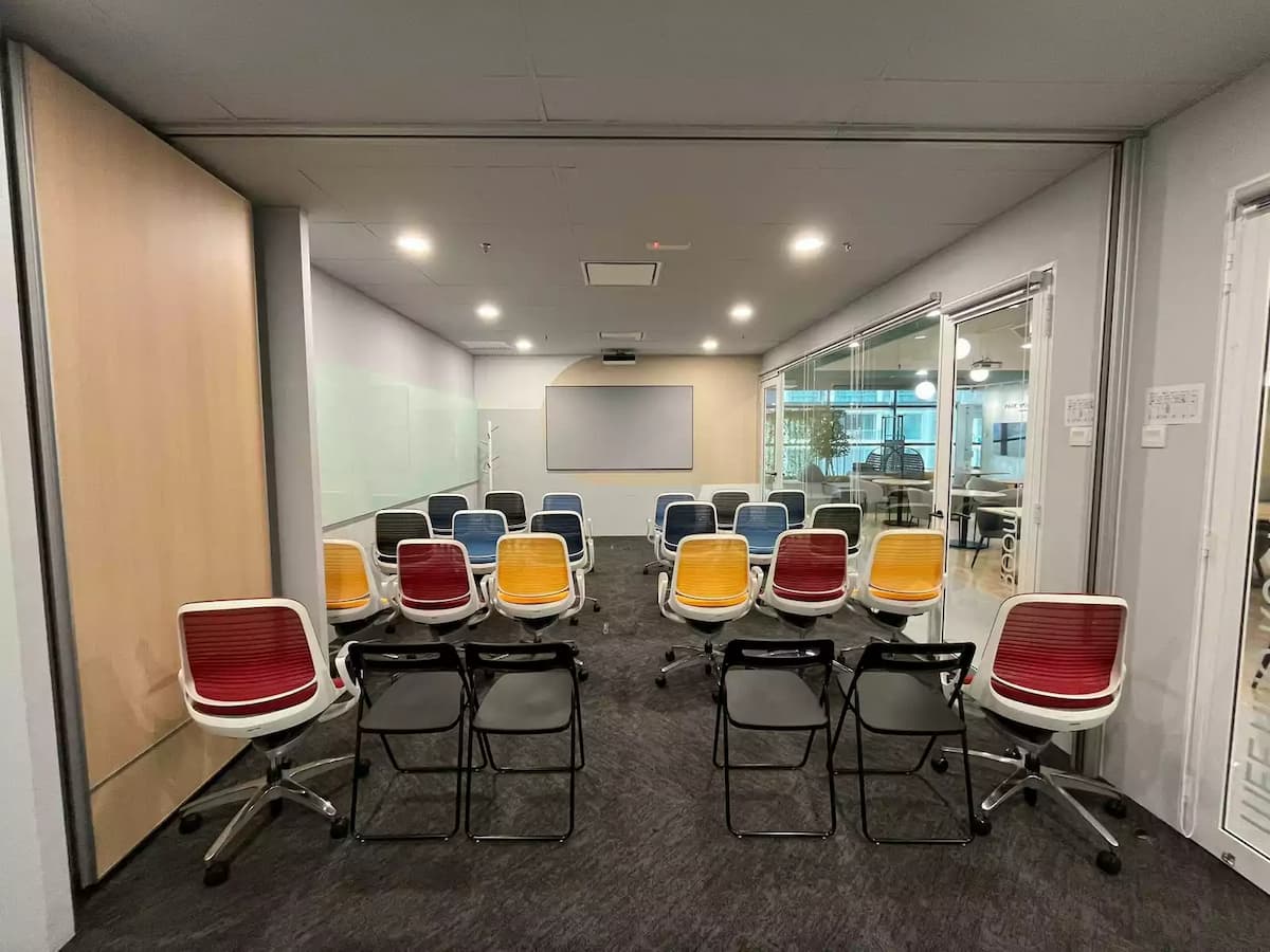 Training-style meeting room with rows of red, yellow, and blue chairs facing a presentation screen, framed by glass walls and sliding partitions.