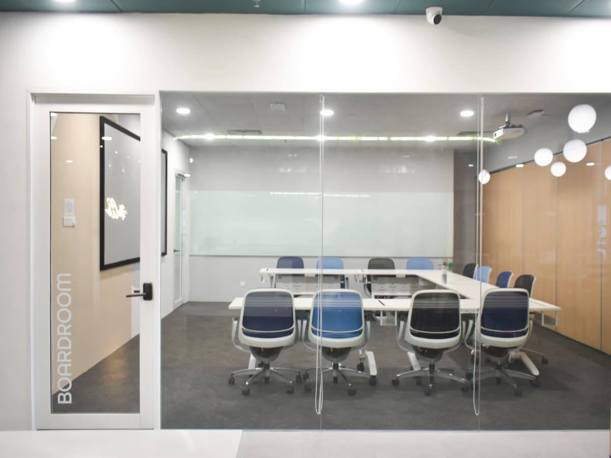 A modern training room with modular white tables arranged in a U-shape, ergonomic chairs, a wall-length glass whiteboard, and a ceiling-mounted projector for presentations.