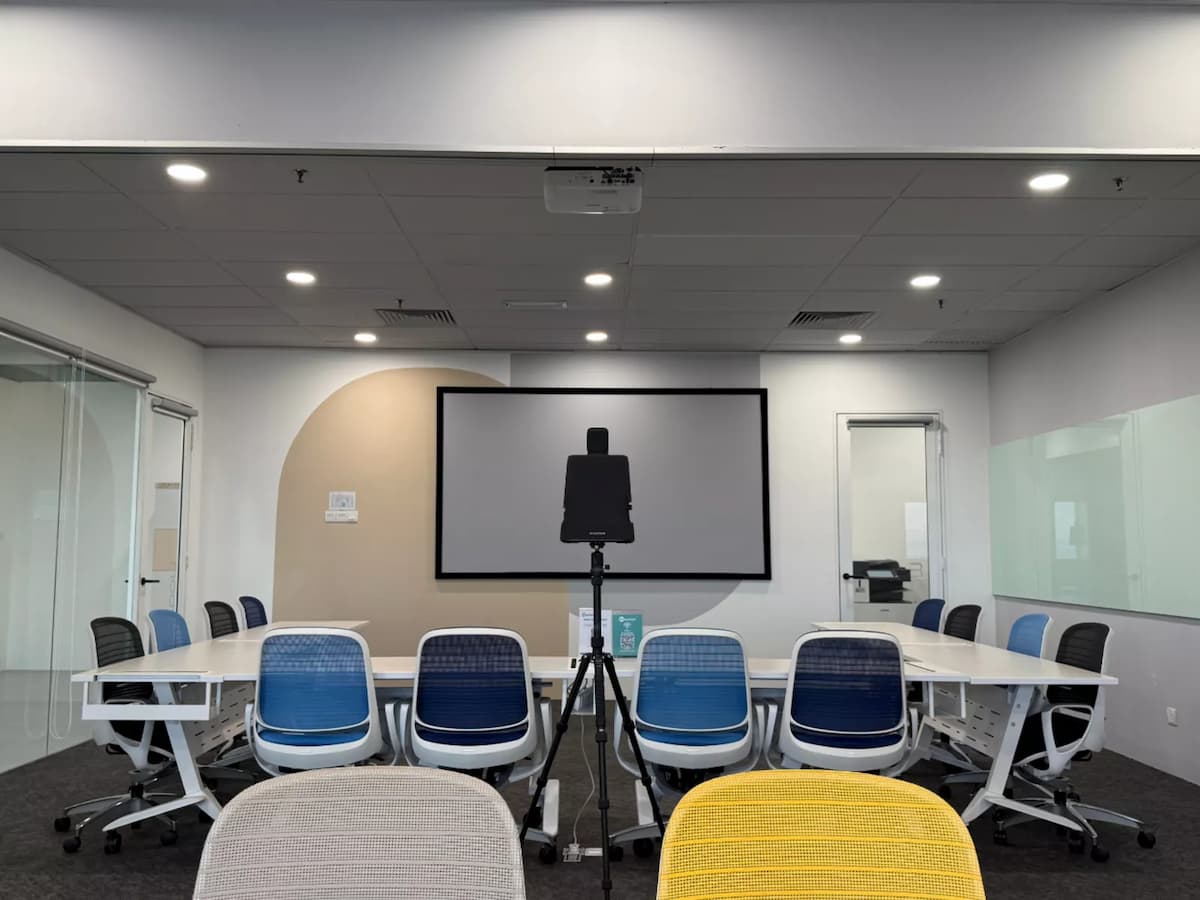A modern training room with modular white tables arranged in a U-shape, ergonomic chairs, a wall-length glass whiteboard, and a ceiling-mounted projector for presentations.