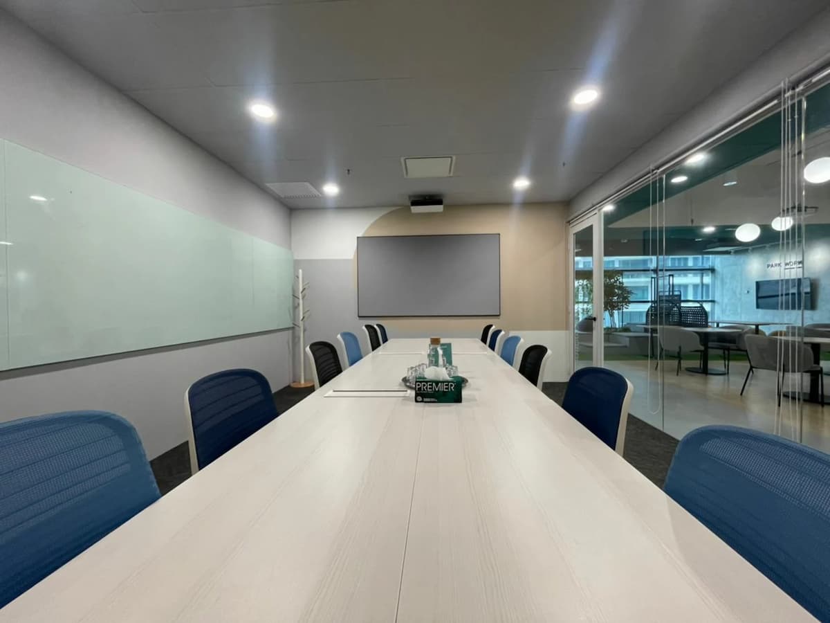 Contemporary meeting room with long conference table, blue and black chairs, glass whiteboard, and presentation screen, viewed from the table’s end.