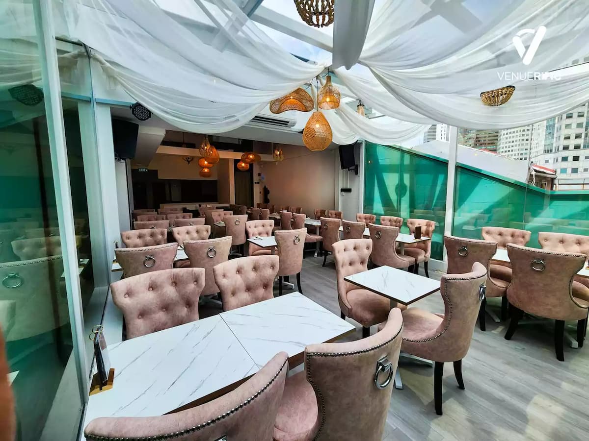 Elegant rooftop event space with blush pink chairs, white draped ceiling, floral backdrop, and gold-accented table settings.