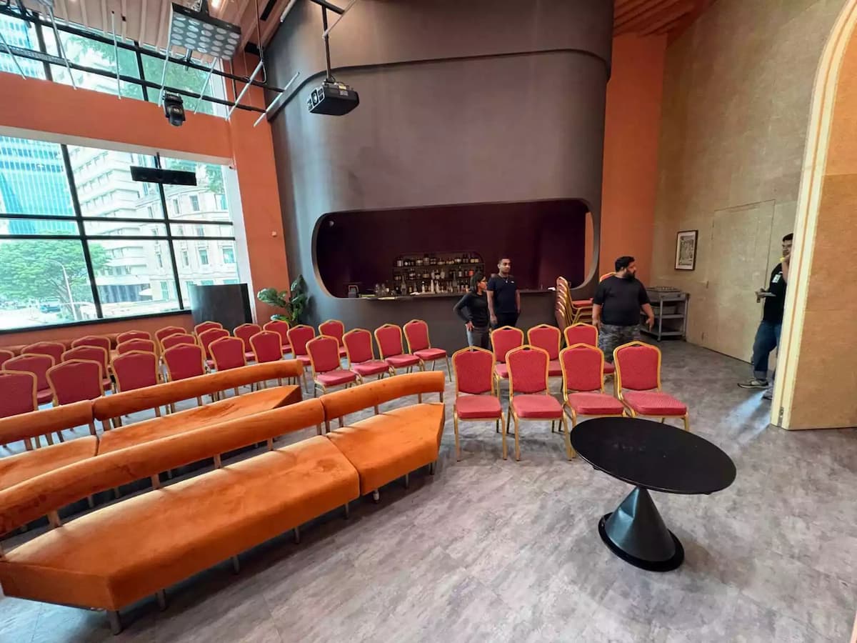Event setup with rows of red chairs, curved sofa seating, high ceiling windows, and a bar counter at the back.