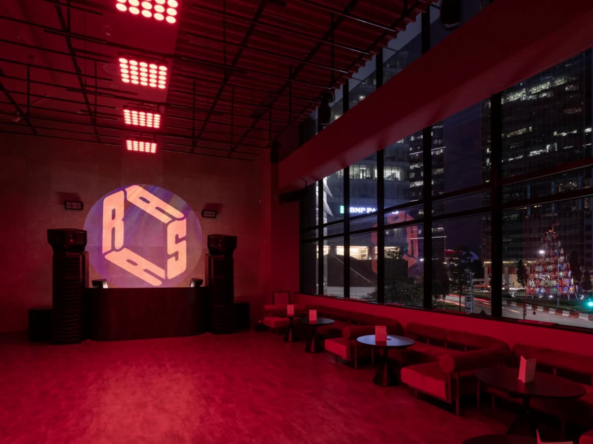 DJ booth with large speakers, red lighting, and city skyline view through floor-to-ceiling windows.