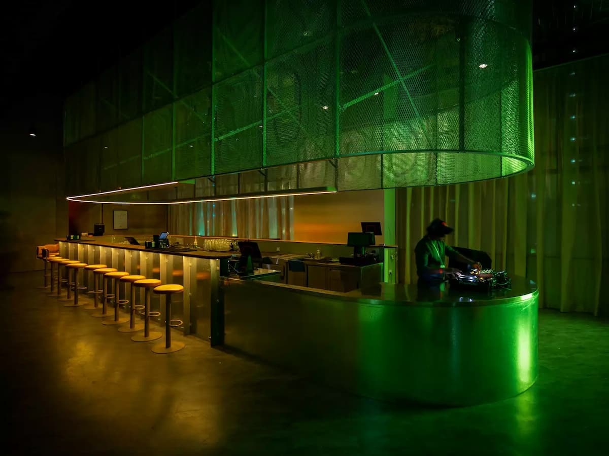 Modern bar with long counter, green ambient lighting, DJ booth, and overhead mesh canopy.