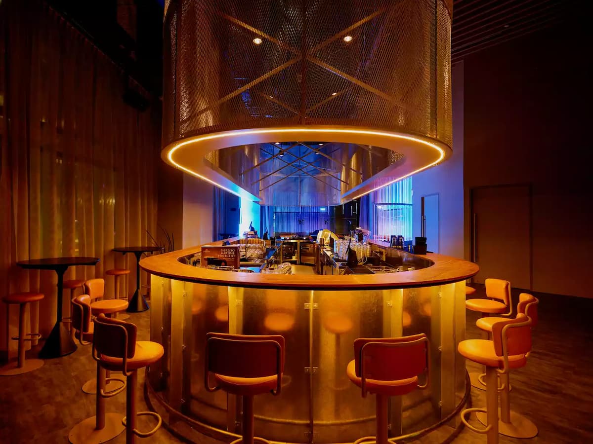 Circular bar counter with warm lighting, overhead mesh canopy, and orange lounge stools.