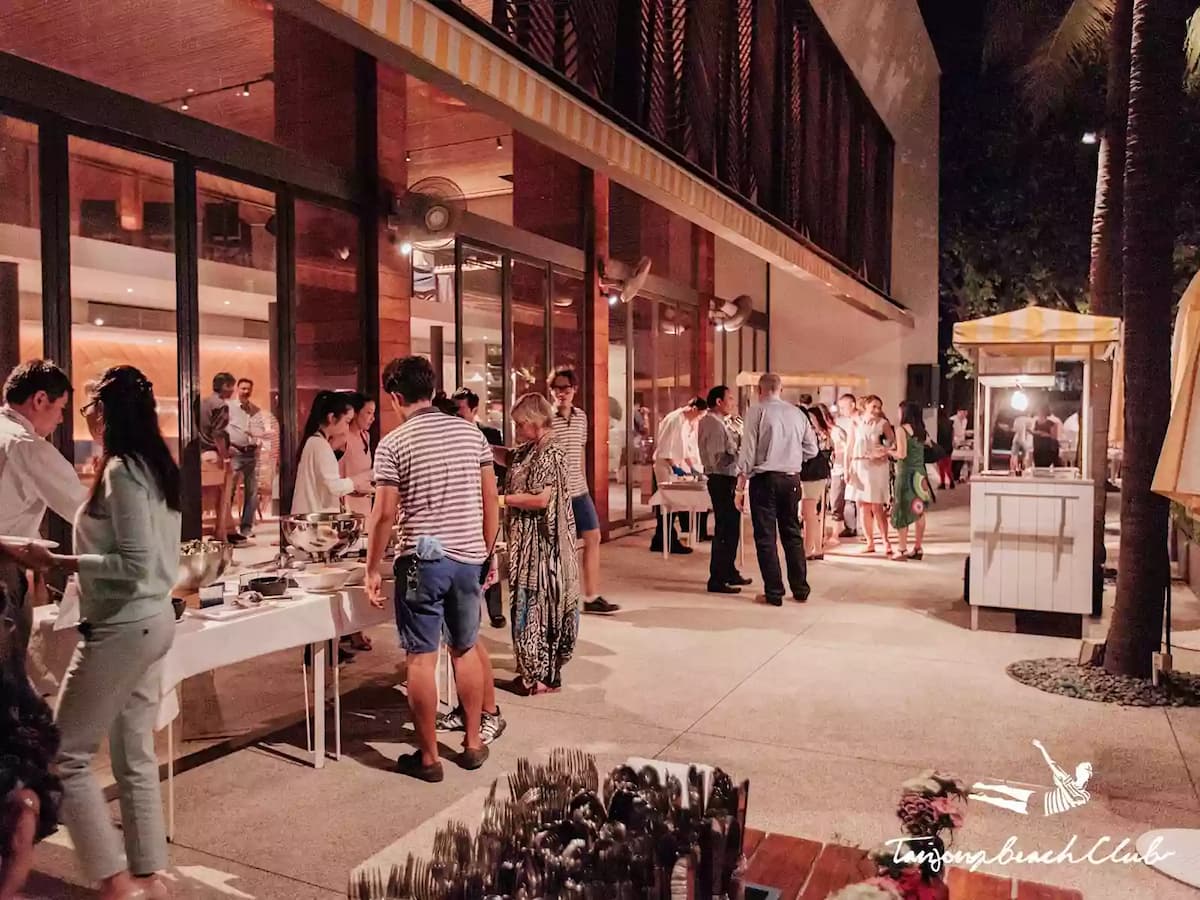 Evening outdoor event at a beach club with guests mingling near buffet tables and lit stalls.