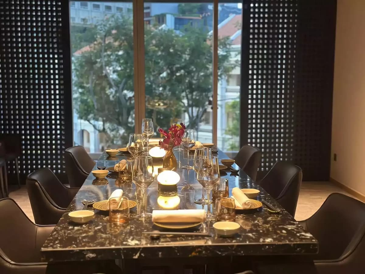 A long, black marble dining table is set for a formal meal with crystal wine glasses, white napkins, and small, glowing table lamps.
