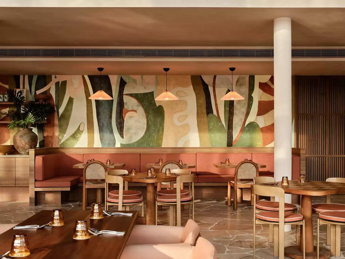 Warm-toned restaurant interior with wooden tables, cushioned seating, and a colorful abstract mural on the wall.