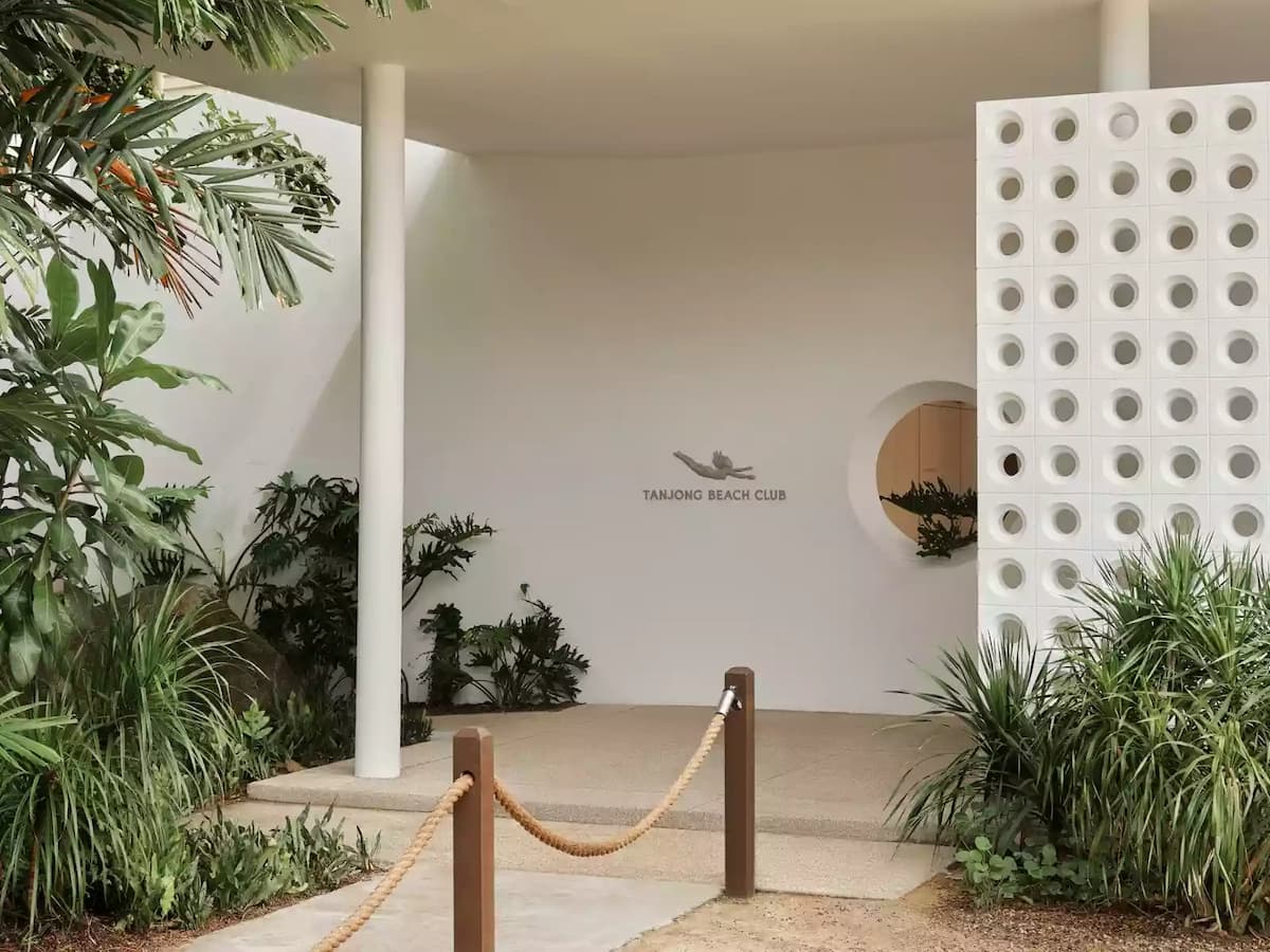 Entrance area with white walls, tropical plants, and “Tanjong Beach Club” signage beside a circular window.