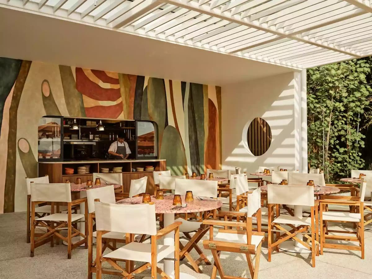 Sunlit outdoor restaurant terrace with wooden chairs, round tables, and a colorful mural backdrop.