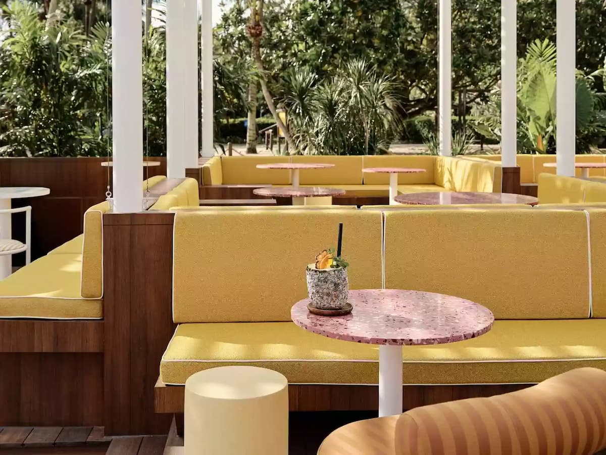 Outdoor café seating with yellow cushioned booths, pink marble tables, and lush greenery in the background.
