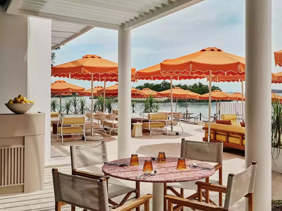 Seaside terrace dining area with round tables, wooden chairs, and orange umbrellas overlooking the water.