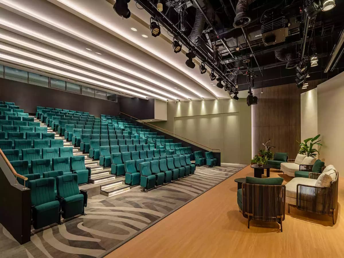 Modern auditorium with tiered seating, stage lounge area, and lighting rig.