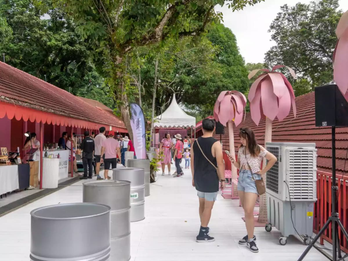 Outdoor market walkway with red-roofed stalls, visitors strolling, and pink decorative installations along the path.