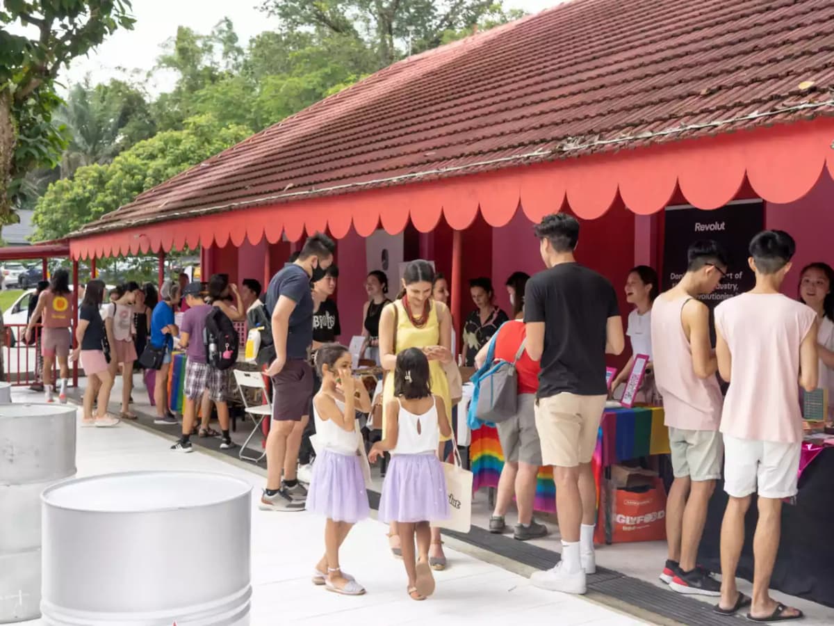Crowd gathered under a red awning at an outdoor pop-up event with booths and families browsing.