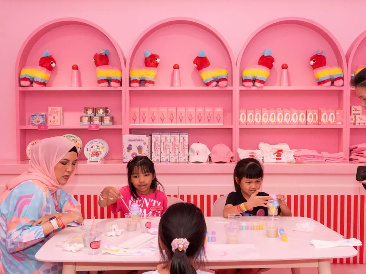 Woman and children crafting at a table in a bright pink retail space with colorful merchandise on arched shelves behind them.