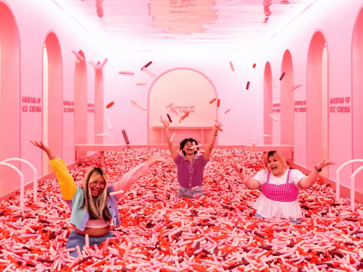 Three people playing in a pink sprinkle pool inside a bright, arched ice cream–themed room.