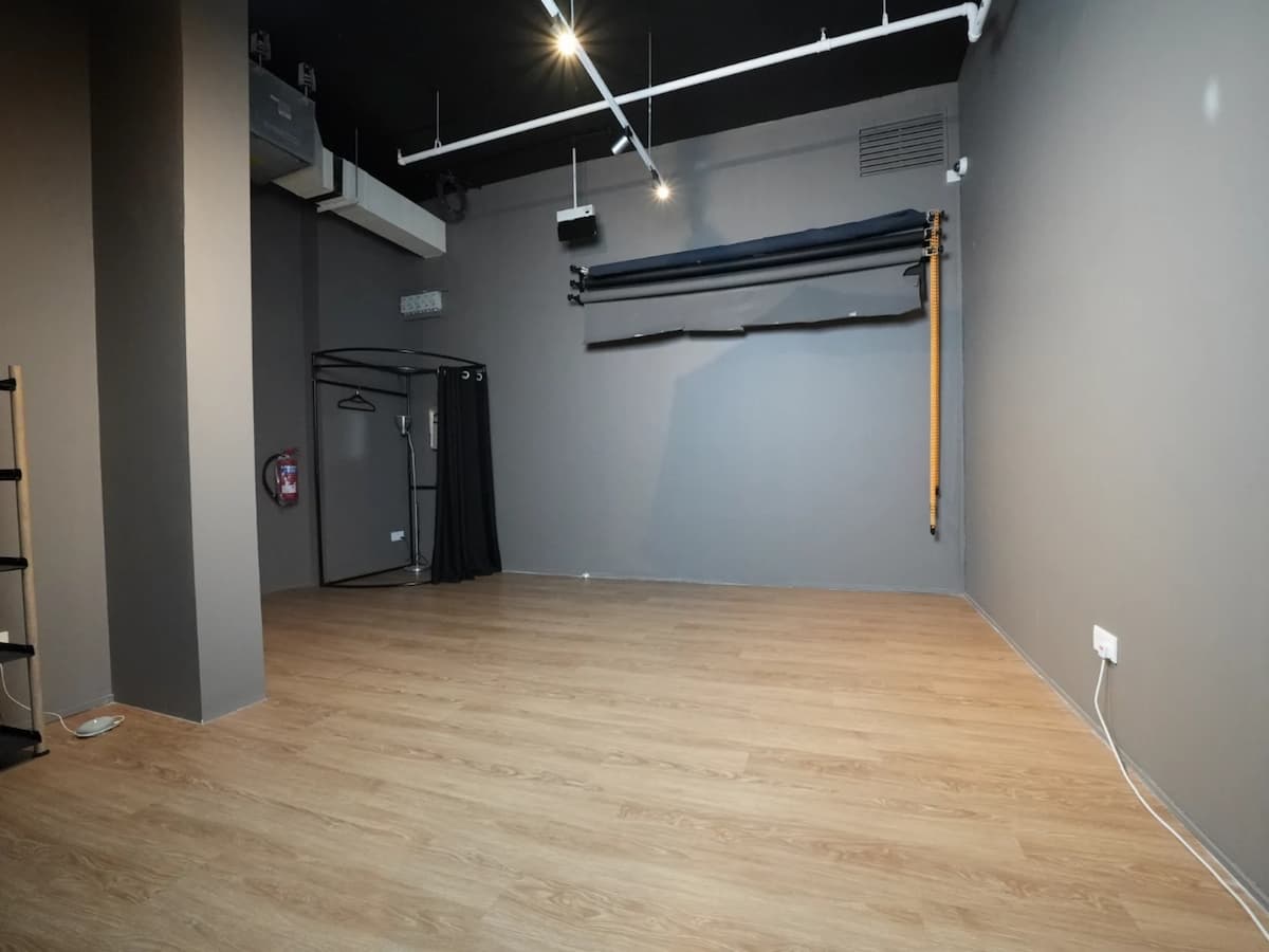 Empty studio space with gray walls, wood flooring, backdrop roll, and a small changing booth in the corner.