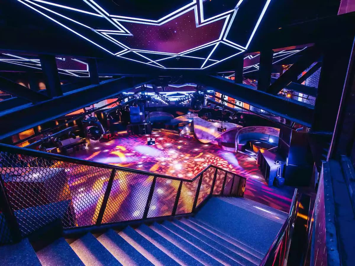 Elevated view of a nightclub dance floor with geometric LED ceiling lights, stairs, and immersive lighting effects.