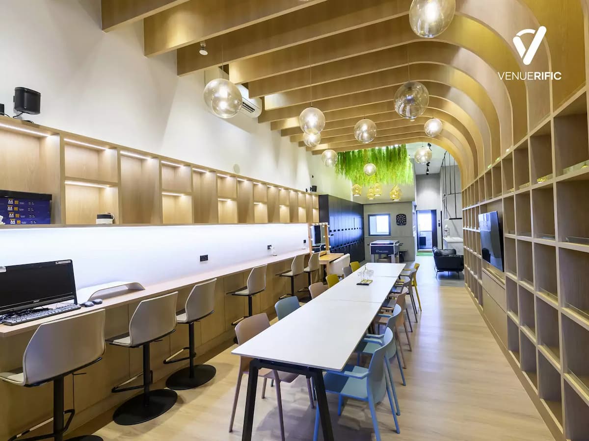 Bright collaborative workspace with long communal tables, wood shelving, globe pendant lights, and greenery accents.