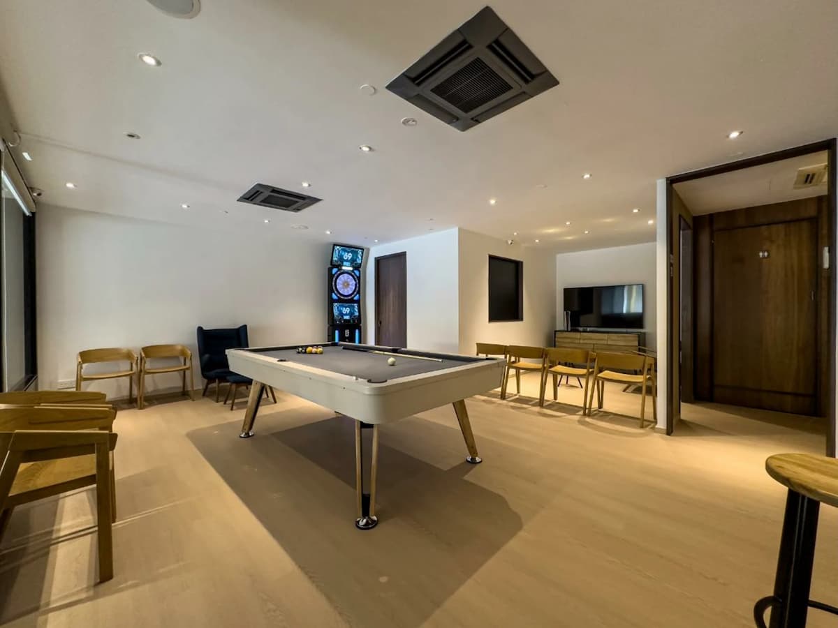 A minimalist recreation room featuring a light-toned pool table, a digital dartboard, and wooden seating against clean white walls.